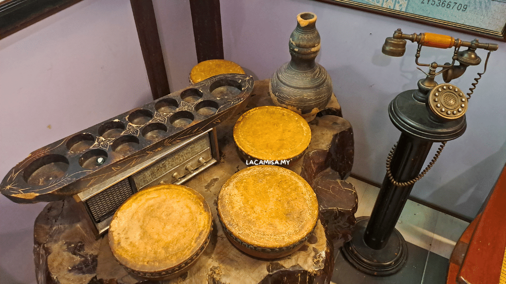 The long wooden board with holes is called 'congkak', a Malay traditional popular game. The three small drums are called 'kompang' a traditional Malay musical instruments.