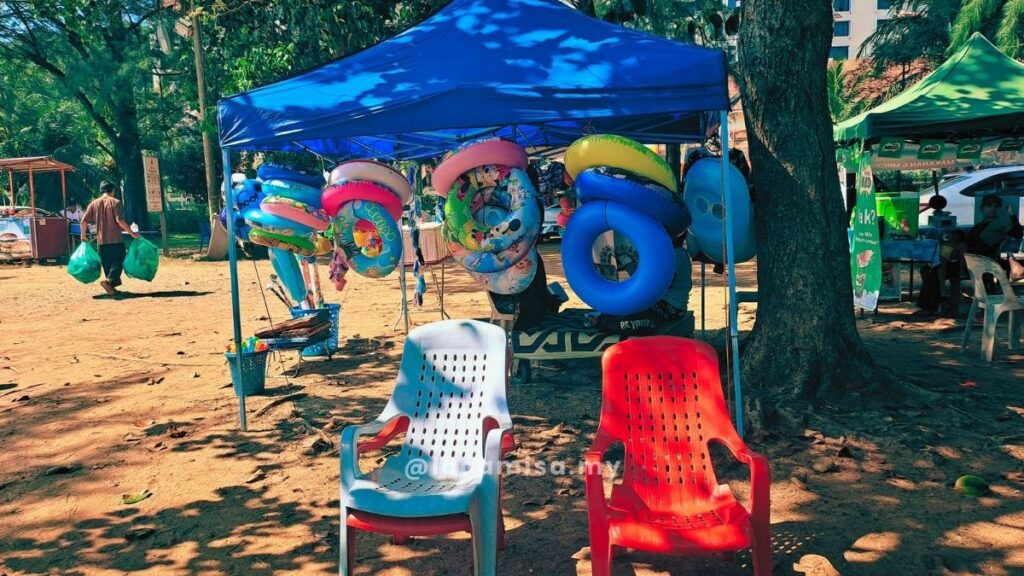 A beach rental stall at Monkey Bay Beach, Port Dickson, featuring colorful inflatable floaties and swim rings hanging under a blue canopy.