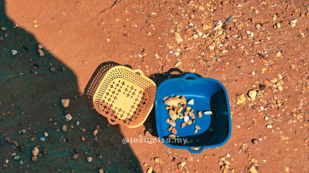 A yellow plastic sieve and a blue plastic basin filled with collected sea shells and pebbles on the sandy shore of Monkey Bay Beach.