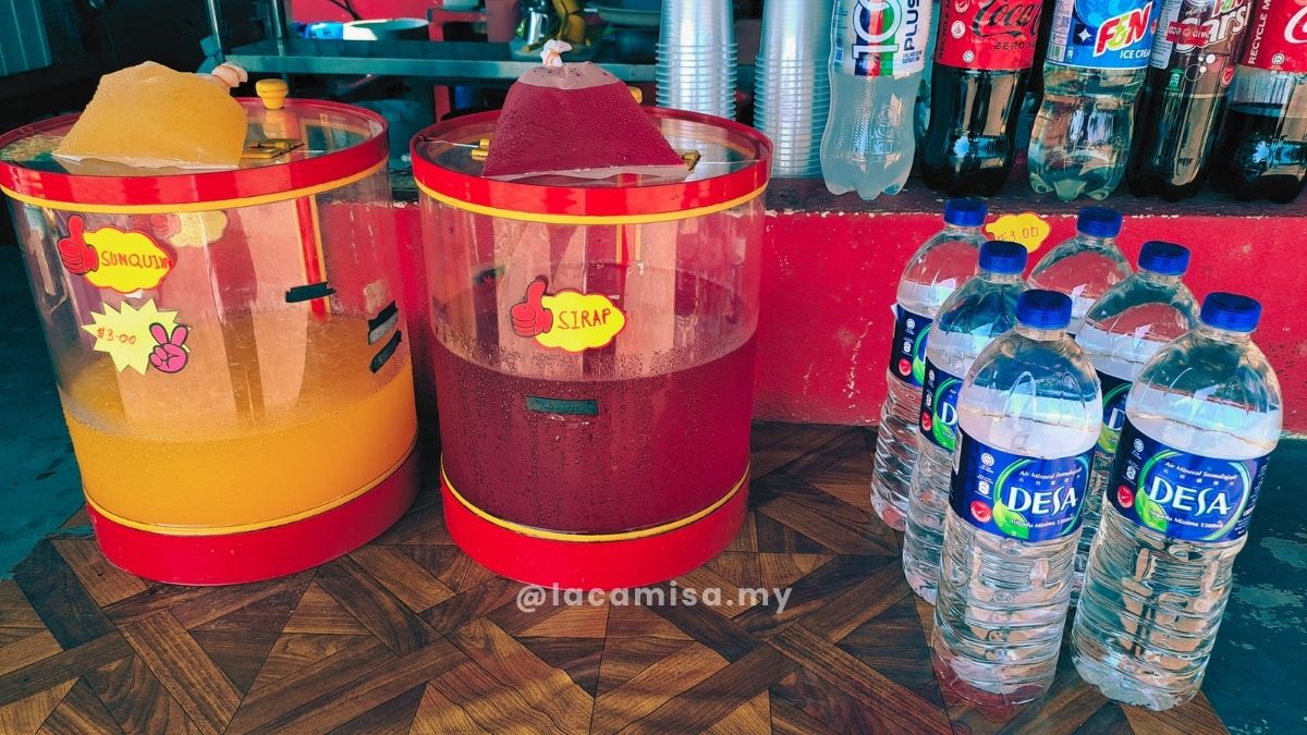 Local beverage stall at Monkey Bay selling Sunquick and Sirap drinks in large dispensers.