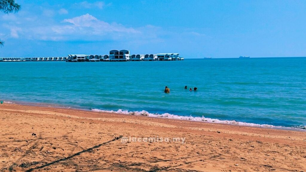 Families and children swimming in the ocean at Monkey Bay Beach with the Lexis Hibiscus Port Dickson resort visible in the background.