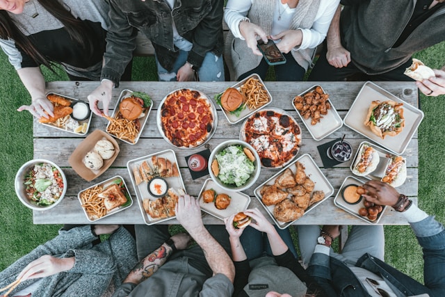 An overhead, bird's-eye view shows a group of casually dressed people gathered around a rustic wooden picnic table outdoors on a green lawn, enjoying a massive communal feast. The table is densely packed with a variety of comfort foods, including two pepperoni pizzas, fried chicken, burgers, fries, fish and chips, tacos, and several bowls of salad. Multiple hands reach into the frame to grab fries, dip appetizers, or hold food, while one person uses a smartphone to capture a photo of the spread.