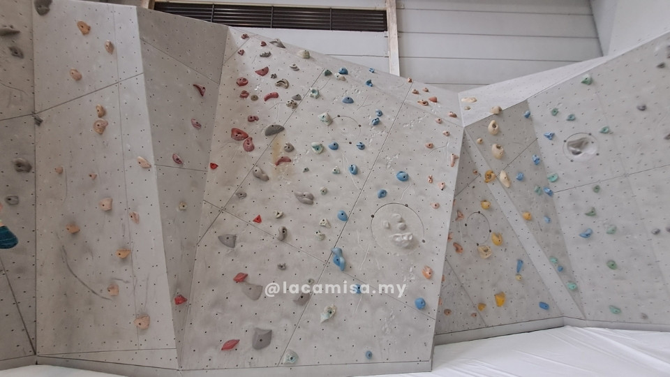 A low-profile bouldering wall at Putrajaya Challenge Park, specifically designed for children and beginners. The wall features a grey, textured surface with various colorful climbing holds and is equipped with a thick, white safety mat at the base to provide a soft landing.