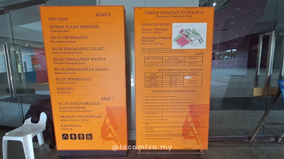 Two orange information boards at the entrance of Putrajaya Challenge Park. The left panel displays the facility directory, listing the Pro Shop, Climbing Hall, and changing rooms on the Ground Floor (Aras G), and the Children’s Climbing Room, Cafeteria, and offices on Level 1 (Aras 1). The right panel outlines the affordable entrance fees at just RM6.00 for adults and RM3.00 for students, alongside a list of equipment rental charges for ropes, shoes, and many more as well as the park's operation hours and house rules.