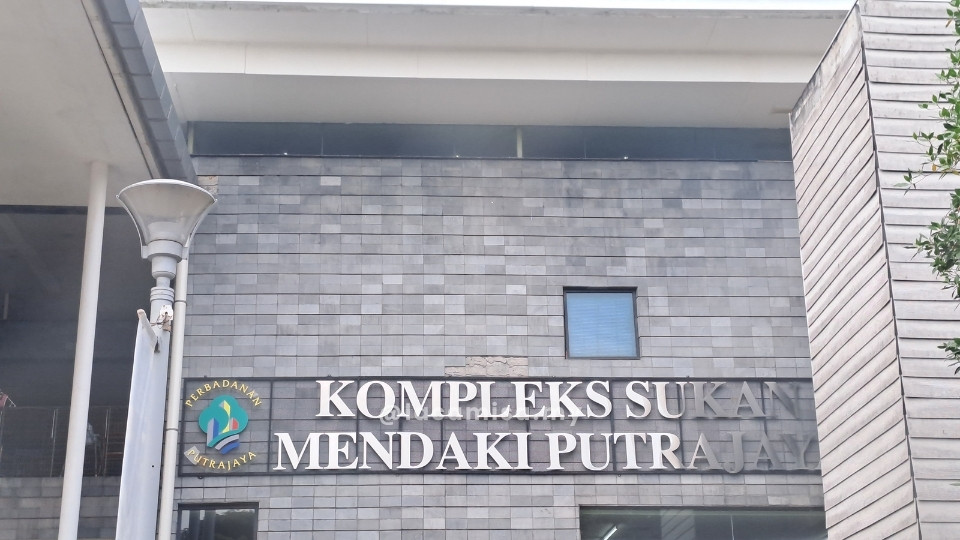 A close-up shot of the exterior entrance wall of the Kompleks Sukan Mendaki Putrajaya (Putrajaya Climbing Sports Complex). The wall is constructed from textured, slate-grey rectangular stone tiles. Mounted on this wall are large, silver metallic letters that spell out the name of the complex in a clean, sans-serif font. To the left of the text is the Perbadanan Putrajaya logo, which features a colorful, stylized graphic enclosed in a gold-bordered circle. The top of the building has a modern white overhang, and a tall, silver outdoor lamp post is visible on the far left, suggesting a well-lit and professional sporting facility.