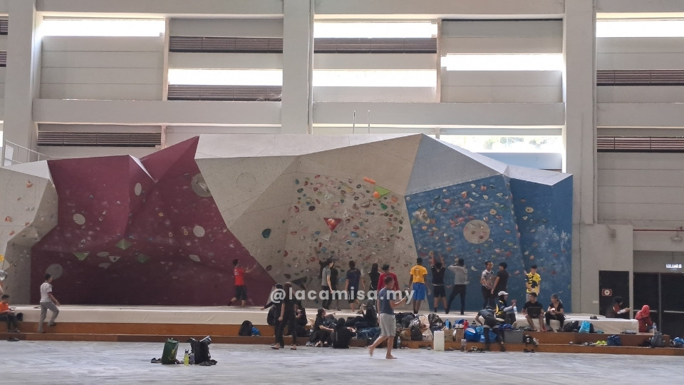 A wide, low-profile bouldering wall at Putrajaya Challenge Park, featuring a multi-angled design in magenta, grey, and blue. The structure is wide but intentionally shorter in height, allowing climbers to practice without ropes. A diverse group of people is seen at the base and on the padded platform, creating vibes where each group is focused on their own goals while actively cheering and supporting one another within the spacious climbing hall.